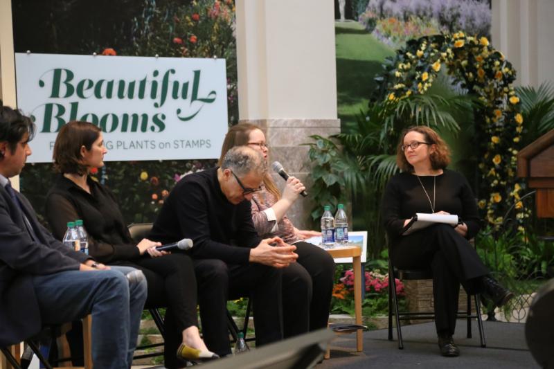 Stage with four seated adults, a white sign behind them to the left and plants to the right