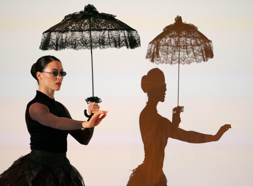 Woman on the left wearing a black dress, sunglasses, & holding a lacy umbrella & her shadow to the right