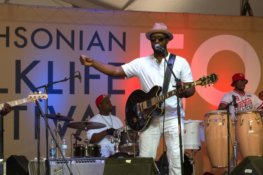 Singer with a guitar stands at a microphone in front of other band members on a stage