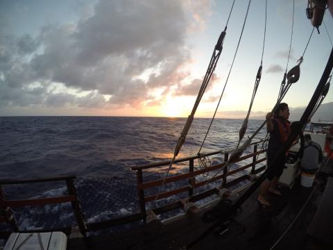 Photograph of woman standing on a sailboat with and ocean and sunrise to left